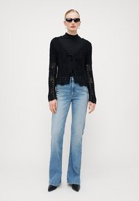 Cardigã de crochê preto com fecho de laço, usado sobre um suéter de gola alta preto. Combinado com uns jeans flare de tom azul claro e uns sapatos de salto agudo pretos.