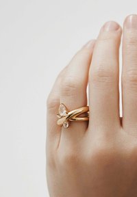 Mano con dos anillos de oro en el dedo anular, cada uno con una piedra preciosa transparente, contra un fondo liso claro.