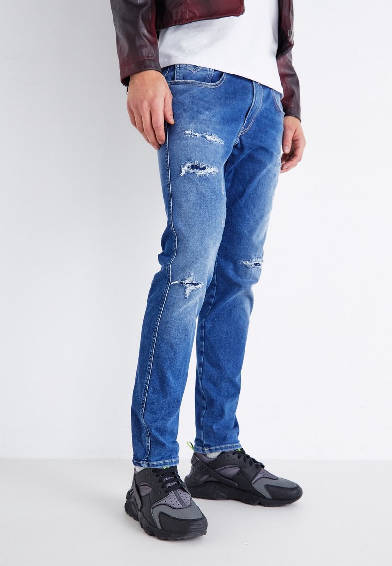 Hombre vistiendo jeans rasgados azules, zapatillas negras y grises, camisa blanca y chaqueta de cuero burdeos contra un fondo blanco.
