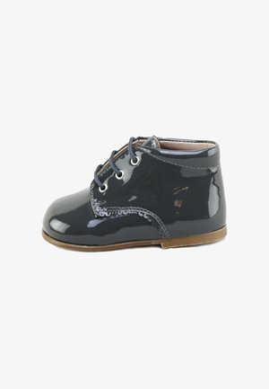 Zapato de charol negro infantil con punta redondeada, diseño con cordones, costuras en contraste y una pequeña suela marrón. Textura suave.