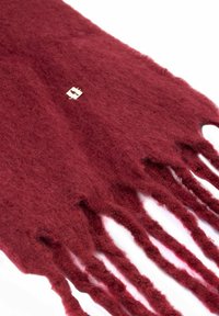 Burgundowy wełniany szal z długimi, frędzlami na krawędziach i małą srebrną metką. Tekstura wydaje się miękka i puszysta.