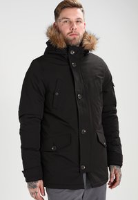 Svart puffy parka-jakke med hette foret med falsk pels, knappelukking, to frontlommer og en glidelåslomme på siden. Myk tekstur.
