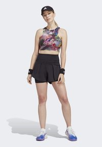 Sport crop top i livlige farver og mønstre, parret med sorte plisserede shorts. Accessories inkluderer sorte håndledsbånd og en kasket.