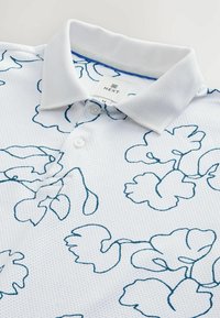 Polo alb cu un material texturat, având un model floral albastru pe margine, cu închidere cu nasturi și guler clasic. Eticheta brandului este vizibilă în interiorul gulerului.