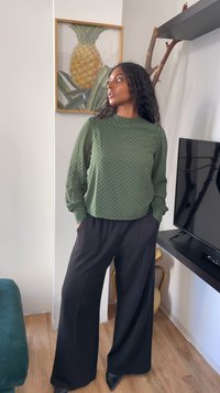 Mulher com blusa texturizada verde e calças largas pretas de pé dentro de casa perto da televisão e de uma obra de arte de abacaxi, olhando para o lado.