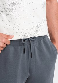 Grå sweatpants med snorelukning og metalstopper, parret med en hvid skjorte med et spredt sort tekstmønster.