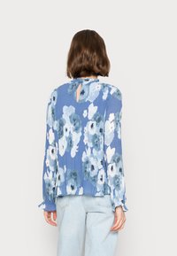 Blusa floral azul clara com mangas longas e textura pregueada; possui um recorte nas costas em forma de chave e punhos elásticos. Combinada com jeans azul claros.