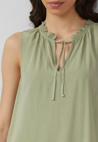 Haut sans manches vert clair avec un col froncé, une découpe en V et une fermeture par lien. Texture lisse avec une coupe décontractée.