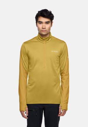 Uomo che indossa una maglia atletica a maniche lunghe color senape con mezza zip e pantaloni neri, in piedi contro uno sfondo chiaro semplice.