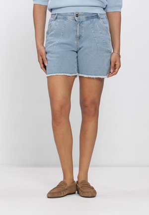 Woman wearing light blue denim shorts with frayed hems, a light blue sweater, and brown moccasin-style shoes, standing against a plain background.