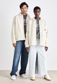 Twee modellen dragen crèmekleurige fleece jassen met knoopsluitingen, gecombineerd met grijze graphic sweatshirts en wijde denim broeken en lichtblauwe rokken.