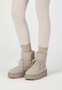 Beige Knöchelstiefel aus einem strukturierten Material, mit dicken Sohlen und Schnürdesign. Kombiniert mit gestrickten beigen Socken und figurbetonten hellen Leggings.