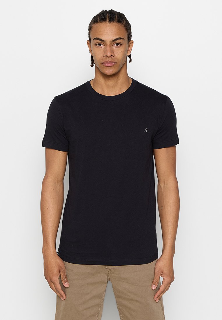 Hombre joven con cabello trenzado, vistiendo una camiseta negra de manga corta y pantalones beis, de pie frente a un fondo blanco.