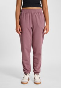 Hellmaulfarbene Jogginghosen mit elastischem Bund, verengten Bündchen und einem dezenten Logodetail am Oberschenkel, kombiniert mit weißen Turnschuhen.