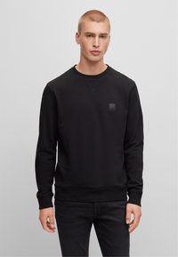Felpa nera realizzata in un tessuto morbido, con scollo rotondo, polsini a coste e una piccola patch con logo nero sul petto. Design semplice.