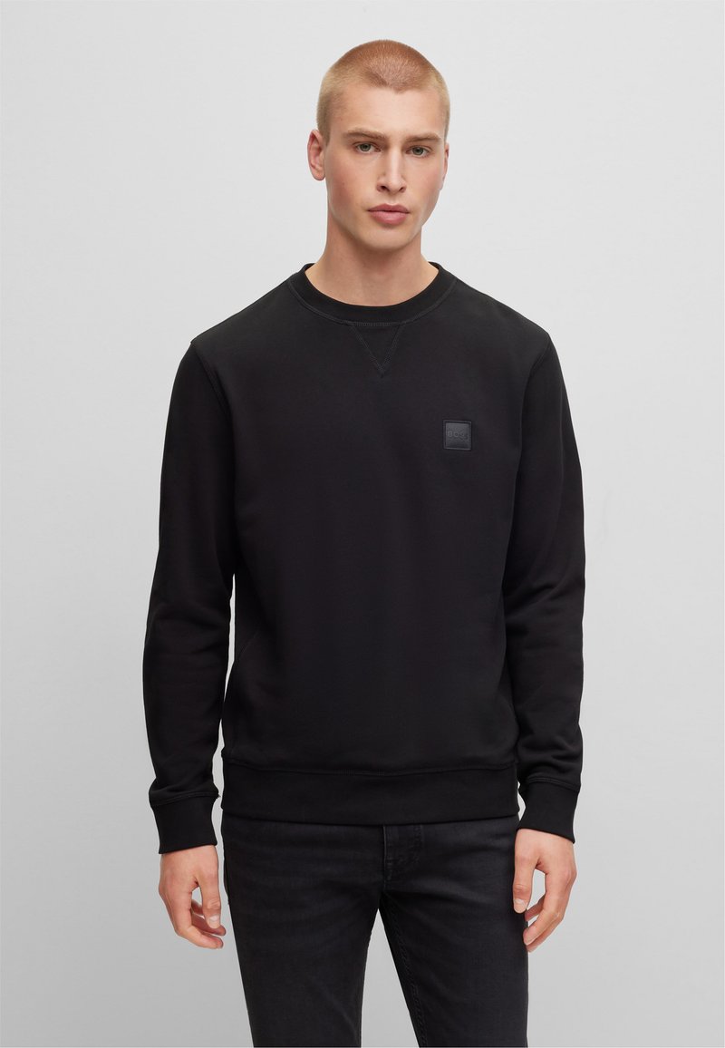 Felpa nera realizzata in un tessuto morbido, con scollo rotondo, polsini a coste e una piccola patch con logo nero sul petto. Design semplice.