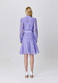 Robe lilas avec de longues manches transparentes et une texture florale, taille cintrée, jupe évasée et escarpins blancs. Conçue avec une ceinture à la taille.