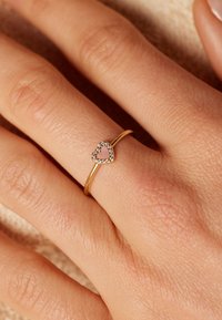 Gouden ring met een hartontwerp met een roze midden en een halo van kleine heldere stenen. Geplaatst op een textuurachtige, lichtgetinte hand.
