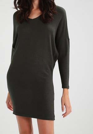 Kobieta w ciemnooliwkowej dzianinowej sukience na długi rękaw, sięgającej do kolan, o luźnym kroju i dekoltem w serek, na jednolitym tle.