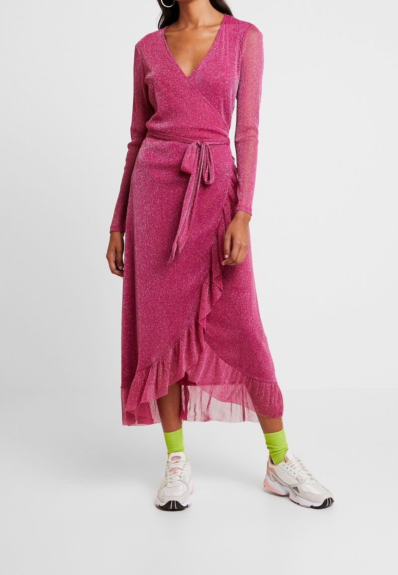 Vestido envolvente rosa, brillante, de manga larga con un dobladillo con volantes y una cinta en la cintura. Combinado con zapatillas blancas y calcetines verde neón. Textura suave.