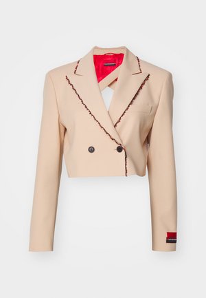 Kivágott bézs zakó, amely szövetből készült, kontrasztos piros béléssel, fekete díszítő pánttal és dupla gombbal záródik.