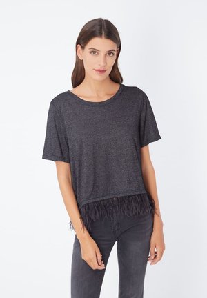 Mujer con cabello castaño viste una camiseta de manga corta gris oscuro con detalle de plumas y jeans gris oscuro, posando frente a un fondo blanco.