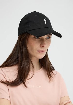 Vrouw met lang bruin haar, die een zwarte baseballpet draagt met een wit polospelerlogo en een lichtroze shirt met de tekst "Polo Club".