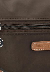 Dettaglio di una borsa in tessuto marrone con una cerniera argentata, etichetta in pelle beige stampata con "handcraft" e superficie testurizzata con dettagli di cucitura.