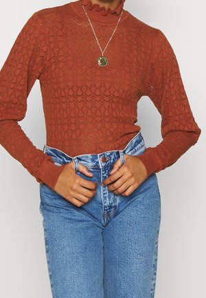 Frau trägt ein rostfarbenes, strukturiertes Langarmshirt und hoch taillierte blaue Jeans, ihre Hände justieren den Bund, dazu eine goldene Anhänger-Halskette.