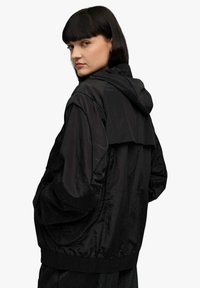 Giacca nera con cappuccio in tessuto leggero, caratterizzata da una vestibilità comoda, polsini a costine e un design posteriore a strati distintivo.