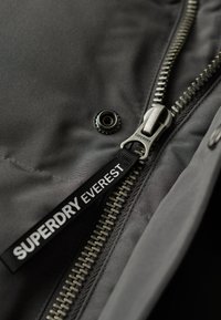 Szary materiałowy żakiet z metalowym zamkiem błyskawicznym, z uchwytem z logo "SUPERDRY EVERREST" oraz detalem z zatrzaskiem. Widok z bliska.