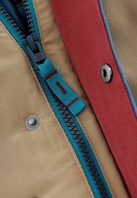 Gros plan sur une veste beige avec une fermeture éclair et un tire-zip bleu sarcelle, une doublure en tissu rouge et des boutons-pression argentés le long du bord.