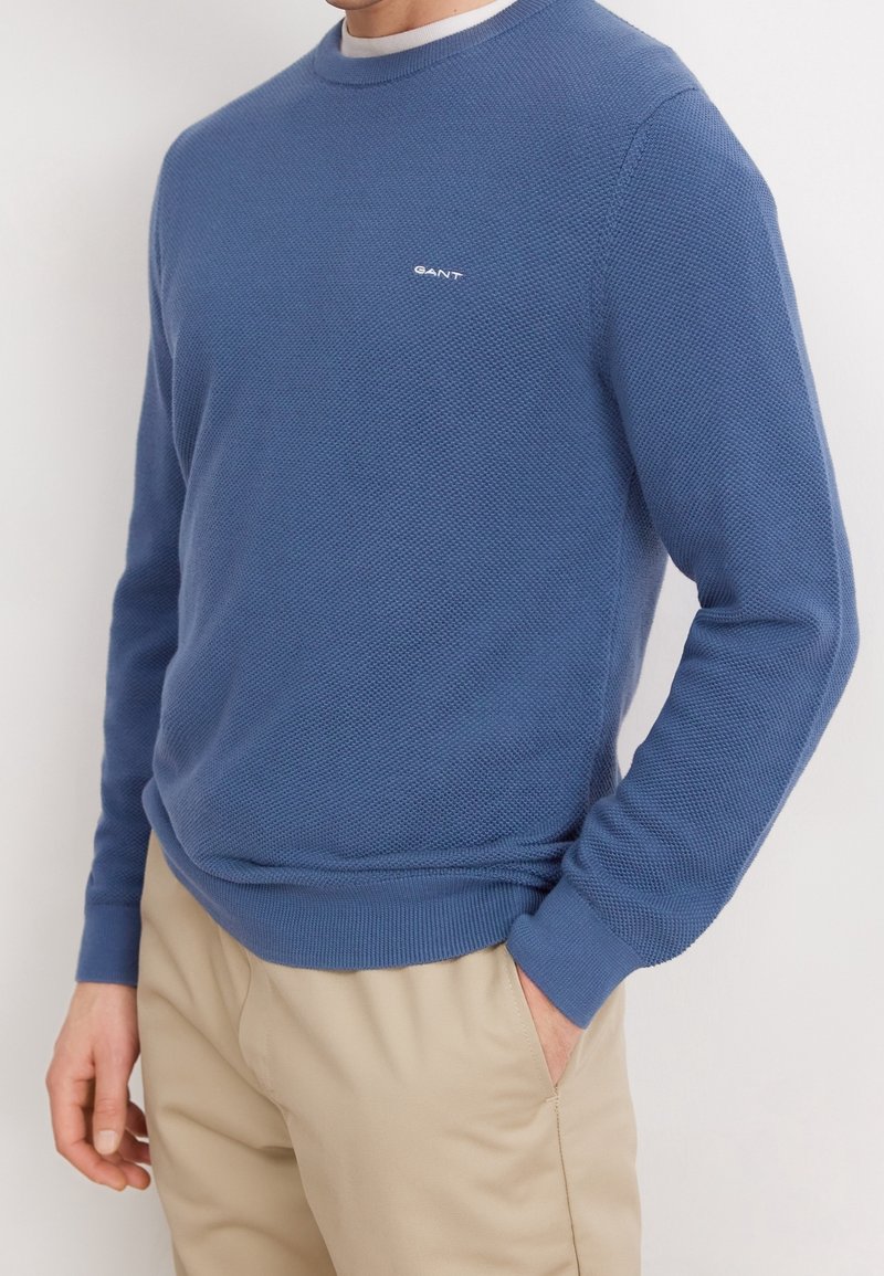 Homme portant un pull à manches longues bleu texturé avec le logo GANT et un pantalon beige, une main dans la poche, debout devant un fond blanc.
