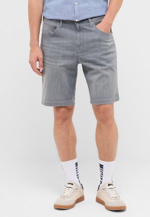 Mann trägt graue Jeans-Shorts, weiße Socken mit blauem Text, beige und weiße Sneakers und ein hellblaues Hemd mit Knöpfen, eine Hand in der Tasche.