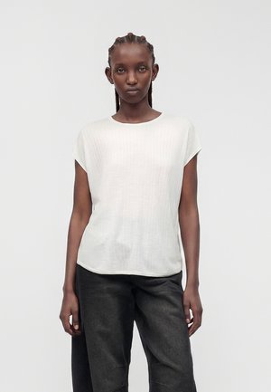 Femme aux cheveux tressés portant un haut blanc texturé à manches courtes et un pantalon noir ample, debout devant un fond blanc uni.