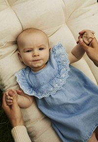 Svijetloplava traper haljina s izvezenim ovratnikom u obliku školjke, ukrašena cvjetnim uzorcima. Beba drži ruku s odraslom osobom na mekanom bež površini.