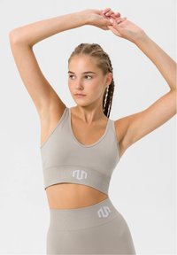 Sujetador deportivo beige con escote en V y banda elástica ancha que presenta un logotipo. Fabricado en una tela suave y elástica con textura acanalada.