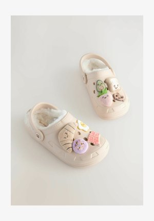 Zoccoli beige con un interno morbido e plush, decorati con vari patch a tema gastronomico, tra cui un avocado, del pane tostato e un uovo sorridente.