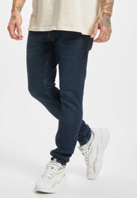 Mann iført mørkeblå smale jeans, hvite joggesko og en lys beige skjorte, stående med det ene benet krysset over det andre.