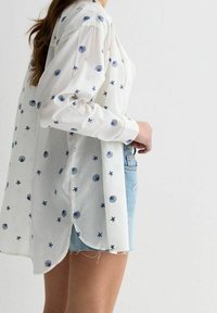 Camicia bianca con motivi a conchiglia blu e stelle marine, con bottoni, maniche lunghe e un orlo leggermente curvo. Abbinata a pantaloni corti di jeans chiari.