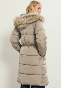 Abrigo acolchado beige con capucha forrada de piel, cintura ajustada y textura acolchada; cuenta con bolsillos laterales y una longitud hasta medio muslo.