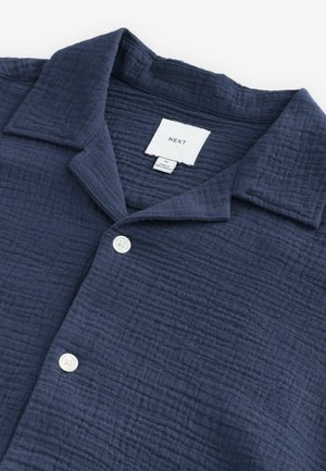 Mornarskoplavi košulja na dugmad izrađena od teksturirane tkanine, s raširenim ovratnikom i jednim bijelim dugmetom sprijeda. Vidljiva oznaka branda.