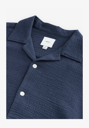 Námořnická modrá košile na knoflíky vyrobená z texturované látky, s rozšířeným límcem a jedním bílým knoflíkem vpředu. Viditelná značka.