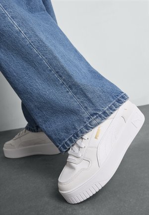Baskets en cuir blanc avec semelles épaisses, arborant un logo doré, associées à un pantalon en jean bleu évasé, présentant un look décontracté.