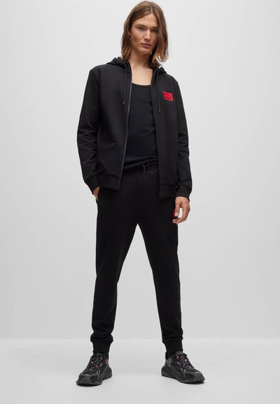 Jeune personne debout portant un sweat à capuche zippé noir avec un patch rouge, un débardeur noir, un jogging noir et des chaussures de sport noires sur fond gris uni.
