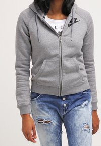 Grå zip-up huvtröja i mjukt material med en figursydd design, kängurufickor och en svart Nike-logga på bröstet. Bärs med slitna jeans.