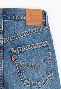 Jeans in denim di blu medio, con un'etichetta in pelle, cuciture arancioni a contrasto e una linguetta rossa sulla tasca posteriore.