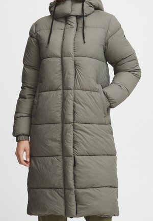 Frau trägt langen olivgrünen, gesteppte Steppmantel mit Kapuze, elastischen Bündchen und Händen in den Taschen vor einfarbigem Hintergrund.