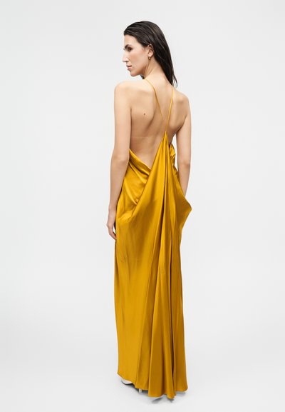 Γυναίκα που φοράει ένα μουσταρδί κίτρινο σατεν φόρεμα με ανοιχτή πλάτη και λεπτές τιράντες, στέκεται με την πλάτη προς το απλό φωτεινό φόντο.