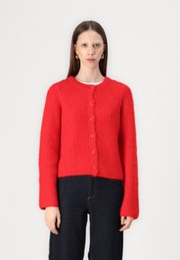 Cardigan rouge en tricot avec un motif texturé, encolure ronde et sept boutons roses sur le devant. Associé à un pantalon en denim noir.